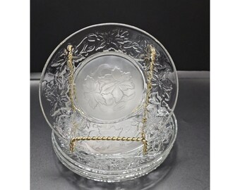 May include: A stack of clear glass plates featuring a raised, textured poinsettia floral pattern around the rim and a frosted, embossed leaf design in the center. The plates are held upright by a gold-toned metal display stand for visibility.