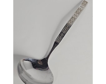 May include: A silver-toned metal ladle featuring a polished, rounded bowl and a long, straight handle. The handle is decorated with a repeating embossed pattern of small flowers and scrolling vine motifs along its entire length.