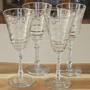 May include: Four clear wine glasses with etched floral designs. The glasses have a classic shape with a round base, a detailed stem, and a flared bowl. The glasses are arranged on a wooden surface.