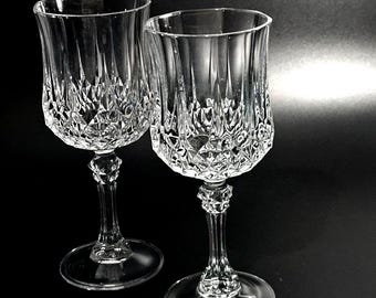 May include: Two clear glass goblets featuring a multifaceted diamond-cut pattern on the lower bowl and a faceted stem design. The glasses have a smooth, flared rim and a circular base, reflecting light through the intricate geometric textures.