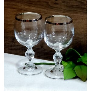 May include: Two clear wine glasses with silver rims and ornate stems. The glasses sit on a white surface with a wooden background. A green leaf is visible in the bottom right corner. The glasses are elegant and suitable for serving wine.