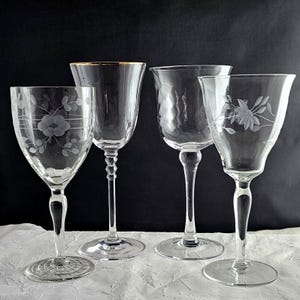 May include: Four clear glass wine glasses with etched floral patterns. One glass has a gold rim. The glasses have long stems and round bases. The background is black and the surface is white.