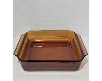 May include: A square baking dish made of transparent amber-colored glass. The dish features a smooth, polished surface with integrated handles on two opposite sides, providing a clear view of the interior base and the four vertical walls.