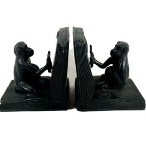 May include: A pair of black bookends, each featuring a monkey figure seated on a book. The monkeys hold a small object, and the bookends are designed to appear as open books. The bookends are made of a dark, possibly cast, material.
