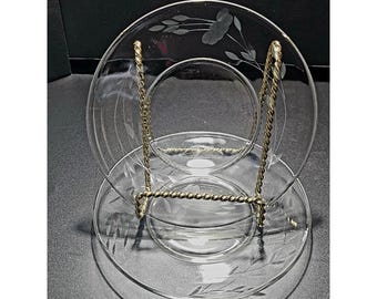May include: A clear glass saucer and matching cup featuring an etched floral pattern with delicate leaves and stems. The set is displayed on a gold-toned, twisted metal stand, showcasing the transparent glass and the fine, etched botanical details.