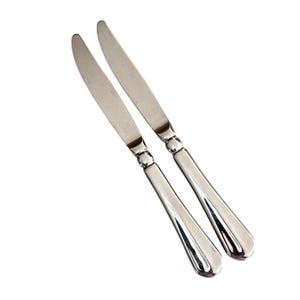 May include: Two stainless steel butter knives with a classic design. Each knife features a rounded blade and a handle with a decorative, slightly curved shape. The knives are set against a white background.