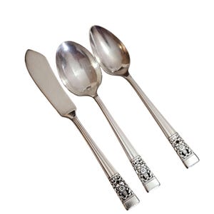 May include: Three pieces of silver cutlery: a butter knife, a spoon, and a teaspoon. The handles have a floral design at the base. The silverware is set against a white background.