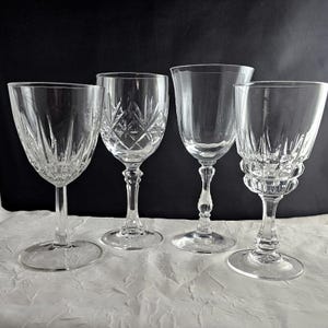 May include: Four clear crystal wine glasses with varying designs. The glasses are displayed on a white surface against a black backdrop. Each glass has a unique stem and bowl shape, ready for use.