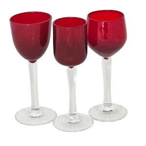 May include: Three wine glasses with ruby red bowls and clear stems and bases. The glasses are arranged in a row, showcasing their elegant design. The bowls have a rounded shape, and the stems are slender.