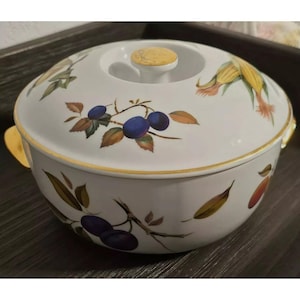 Może przedstawiać: Biała ceramiczna zapiekanka z pokrywką, z kwiatowym wzorem z niebieskimi śliwkami, żółtą kukurydzą i zielonymi liśćmi. Zapiekanka ma złotą obwódkę.