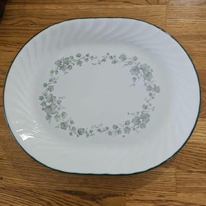 May include: White oval serving platter with a green rim and a floral design in the centre. The floral design features light green and grey leaves and flowers. The platter has a swirled edge.