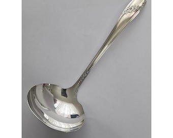 May include: A silver-toned metal soup ladle with a polished, reflective bowl and a long, slender handle. The handle features an embossed floral pattern near the top and a subtle decorative scroll design extending down the neck of the utensil.