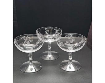 May include: Three clear glass coupe glasses featuring a delicate etched floral pattern along the rim. Each glass has a wide, shallow bowl supported by a slender stem with a decorative knop detail and a circular base for stability.
