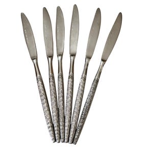 May include: Set of six stainless steel dinner knives. Each knife features a silver blade and a decorative handle with a repeating starburst pattern. The knives are arranged in a fan shape against a white background.