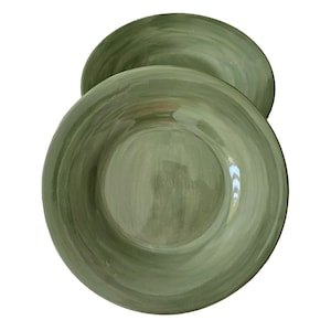 May include: Two green ceramic plates with a wide rim. The plates have a hand-painted appearance with visible brushstrokes, creating a textured, artistic look. The color is a muted, earthy green, suitable for serving food.