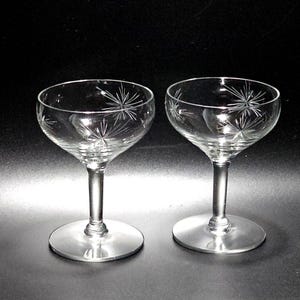 May include: Two clear, stemmed champagne glasses with a starburst design etched into the bowl. The glasses have a round base and a slender stem. The starburst pattern adds a touch of elegance.