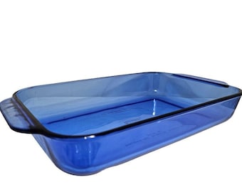 May include: A rectangular baking dish made of translucent cobalt blue glass. The dish features integrated handles on both short ends and has the brand name Pyrex embossed in the center of the interior base, along with other faint text markings.