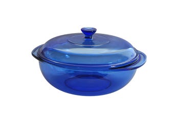 May include: A round, translucent cobalt blue glass casserole dish with two integrated side handles and a matching circular glass lid featuring a central knob handle. The vessel has a smooth, polished finish and a deep, bowl-like base structure.