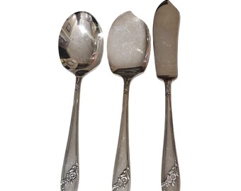 May include: A set of three silver-toned metal utensils consisting of a rounded spoon, a wider oval-shaped spoon, and a flat-edged spreader knife. Each handle features a decorative embossed floral pattern at the base with a polished metallic finish.