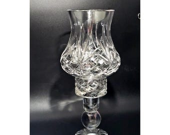 May include: A clear glass candle holder featuring a flared, cut-glass hurricane shade with a starburst and diamond pattern. The base consists of a spherical glass node atop a circular, scalloped foot, providing a stable foundation for the piece.