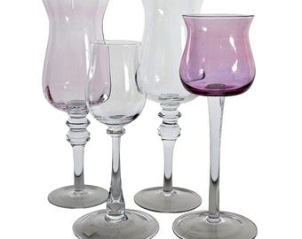 May include: Four stemmed glasses in varying shapes and sizes. Two glasses are clear, one is a light purple, and one is a deeper purple. All have a clear stem and a round base. The glasses are arranged on a white background.