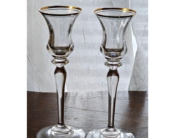 May include: A pair of clear glass candle holders featuring a flared top rim accented with a thin gold band. Each holder has a slender stem that transitions into a wide, circular base, providing a stable foundation for holding taper candles.