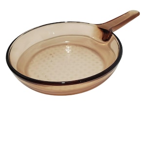 May include: A clear, amber-colored glass skillet with a handle. The skillet has a round shape with a dark brown rim and a textured bottom. The handle is an extension of the skillet's body.