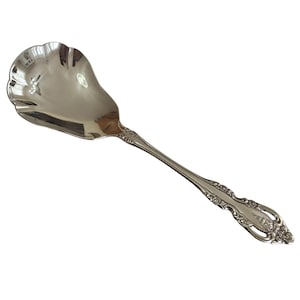 May include: A silver serving spoon with a decorative handle and a scalloped bowl. The spoon has an ornate design along the handle and a polished finish, suitable for serving. The spoon is made of metal.