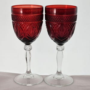 May include: Two matching wine glasses featuring deep red bowls with a textured diamond pattern and embossed leaf motifs. Each glass has a clear, smooth stem with a small decorative faceted knob near the top and a wide, circular clear glass base.