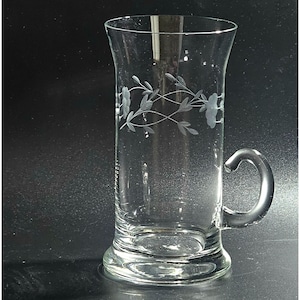 May include: Clear glass mug with a handle and floral etched design. The mug has a flared top and a rounded base. The handle is a curved loop. The etched design features flowers and vines. The mug is suitable for beverages.