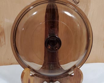 May include: A circular, transparent amber-colored glass lid featuring a smooth, slightly domed surface and a centered, integrated knob handle. The lid has a distinct rim designed to fit securely onto a corresponding cooking vessel or container.