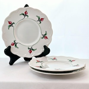 May include: Three white ceramic saucer plates with a green and red floral design. The plates have a scalloped edge and a raised design.