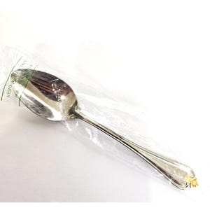 May include: A silver spoon with a decorative handle, still in its original plastic packaging. The spoon has a shiny, reflective surface and a gold-colored accent at the end of the handle. The packaging has the text "REED & BARTON" printed on it.