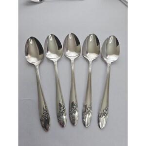 May include: A set of five silver-toned metal spoons arranged in a row. Each spoon features a polished oval bowl and a slender handle decorated with a detailed embossed floral pattern at the base, showcasing intricate rose motifs on the handle ends.