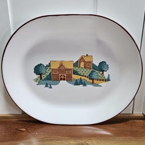 May include: White oval serving platter with a maroon rim. The center features a landscape scene with two yellow and brown buildings, trees, and a fence. The design is in shades of blue, green, and yellow.