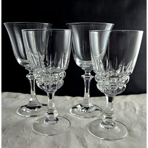 May include: Four clear crystal wine glasses. Each glass has a detailed, textured design on the base and a slender stem. The flared bowls are elegant, ideal for serving wine. The glasses are arranged on a white surface against a black background.
