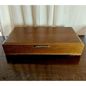 May include: A rectangular wooden box with a medium brown wood grain finish. The box features a flat lid and a centered, horizontal brass-toned metal handle on the front panel. The construction includes a stepped base design along the bottom edge.