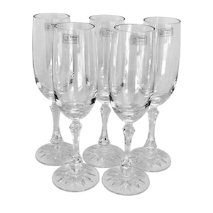 May include: A set of five clear crystal champagne flutes. Each glass has a long stem, a decorative base, and a slender, elongated bowl. The glasses have a label near the top. The flutes are arranged in a circular pattern.
