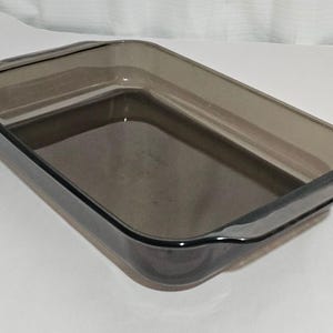 May include: A rectangular, smoky-gray glass baking dish with curved edges and handles. The dish is empty and sits on a white surface. The glass has a slight tint, and the overall design is simple and functional.