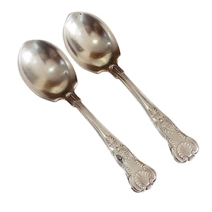 May include: Two antique silver spoons with ornate floral and shell designs on the handles. The spoons have a polished, reflective surface, and are set against a white background. The spoons are likely for serving or dining.