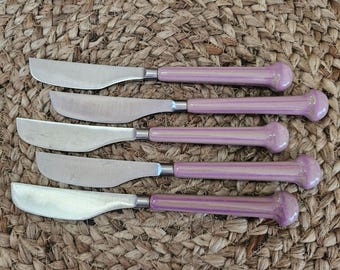 May include: A set of five table knives featuring metallic silver blades and smooth, glossy, light purple ceramic handles with a rounded, bulbous end. Each knife has a consistent design with a curved blade edge and a polished finish on the handle.