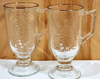 May include: Two clear glass mugs with a footed base and a handle, each featuring an embossed pine tree design on the front and a thin gold rim around the top edge. The mugs are transparent with a smooth texture and a sturdy, elevated stem design.