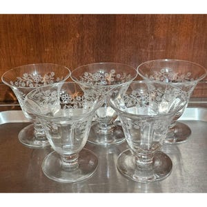 May include: Set of five clear glass dessert dishes. Each dish features a round base, a stem, and a flared bowl. Delicate floral patterns are etched around the bowl's rim. The dishes are arranged on a silver tray.