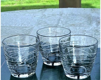 May include: Three clear glass tumblers featuring a series of thin, white, horizontal wavy lines etched around the exterior of each vessel. The glasses have a rounded, slightly tapered shape with a smooth rim and a thick, solid glass base.