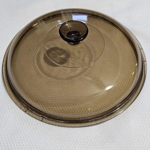 May include: A clear glass lid with a brown rim and a brown knob on top. The lid is designed for a pot or pan.