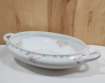 May include: An oval porcelain serving dish with two side handles, featuring a white base decorated with delicate pink rose clusters and green foliage patterns. The rim and handles are accented with thin gold trim, creating a refined appearance.