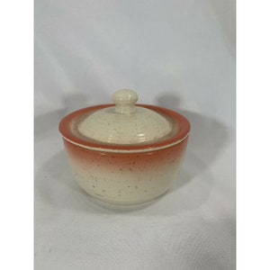 May include: A small, white ceramic jar with a lid. The jar has a speckled glaze and a red band around the top. The lid has a knob on top.