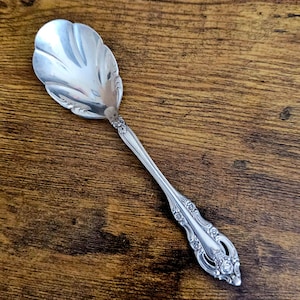 May include: A silver serving spoon with a scalloped bowl and ornate floral detailing on the handle. The spoon is made of a shiny, reflective metal and rests on a dark wooden surface.