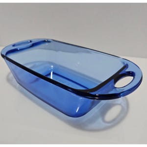 May include: A rectangular baking dish made of translucent cobalt blue glass. The dish features rounded corners and two integrated handles on the shorter ends, each containing a circular cutout for gripping. The glass is smooth and clear.
