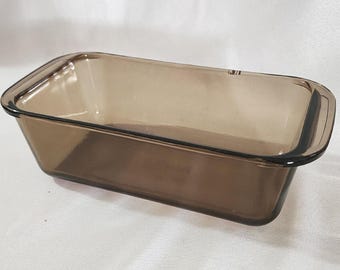 May include: A rectangular loaf pan made of translucent, smoke-colored glass. The baking dish features smooth, rounded corners, a wide rim for easy handling, and deep, straight sides, providing a clear view through the tinted material.